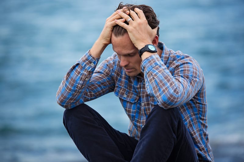 man in blue and brown plaid with both hands in his hair sitting near a body of water