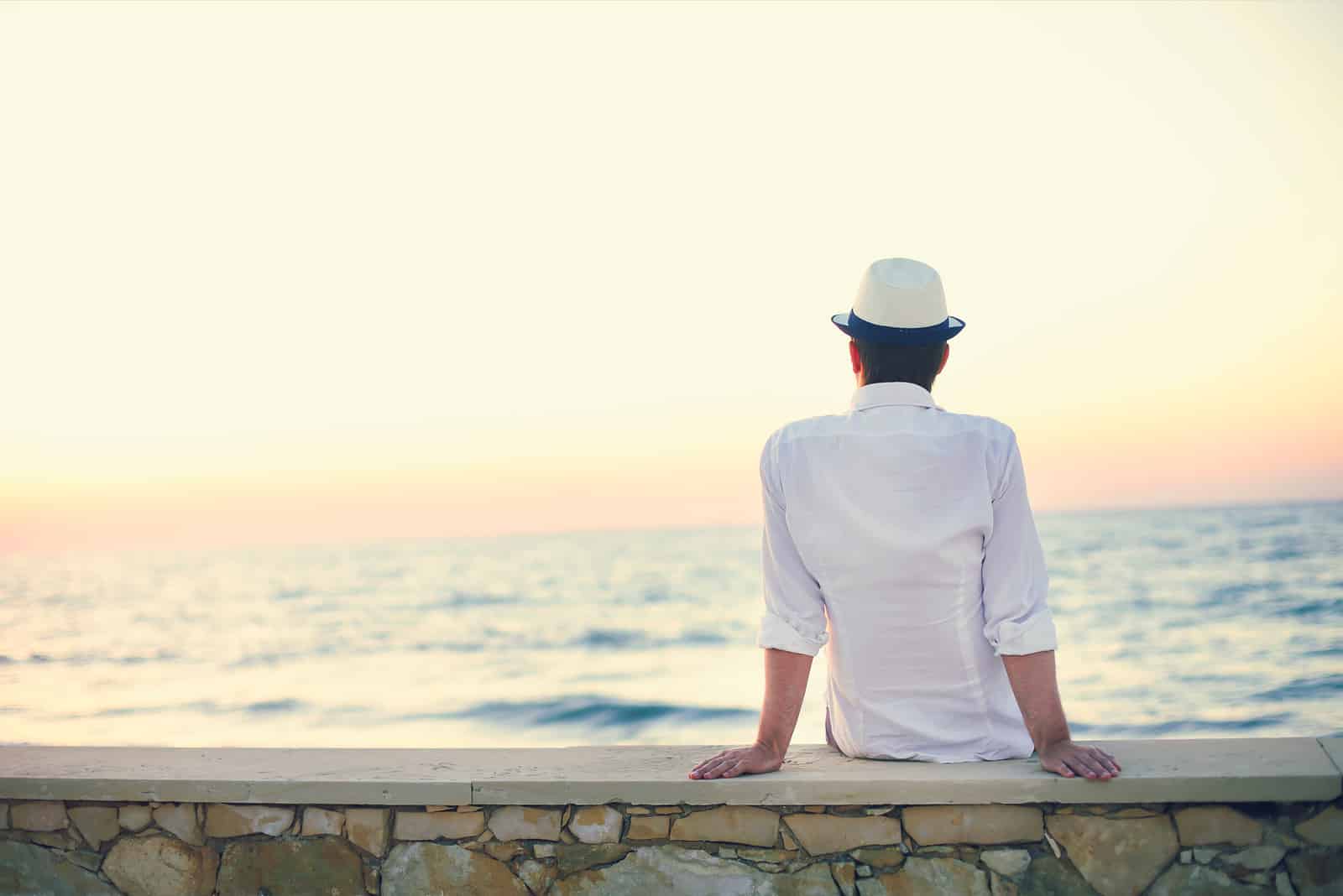 a man with a white hat on his head sits on the wall and looks out to sea